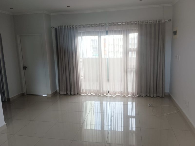 A bright and spacious living area featuring polished floor tiles, recessed ceiling lighting, and large sliding glass doors leading to a balcony.