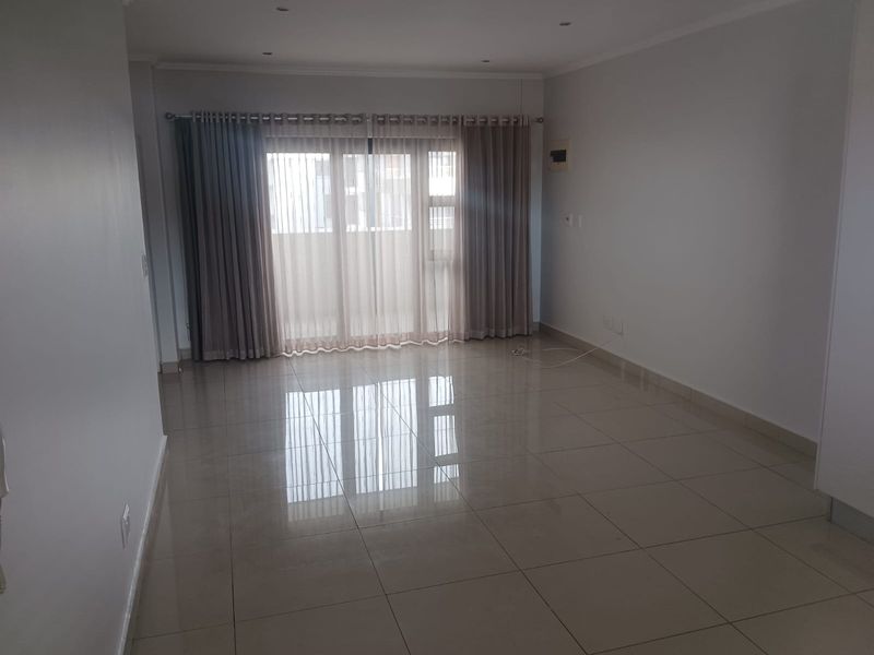 A spacious, empty living area featuring polished porcelain floor tiles, recessed ceiling lighting, and large sliding glass doors leading to a balcony.