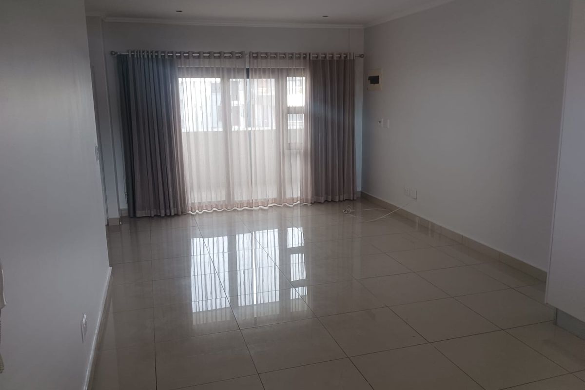 A spacious, empty living area featuring polished porcelain floor tiles, recessed ceiling lighting, and large sliding glass doors leading to a balcony.