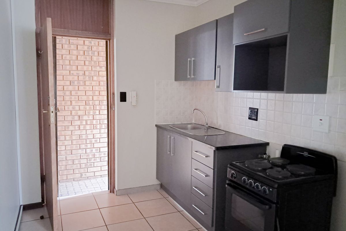 A compact kitchen featuring grey cabinetry, tiled backsplash, ceramic flooring, and a freestanding stove, with direct access to an exterior brick-walled area.