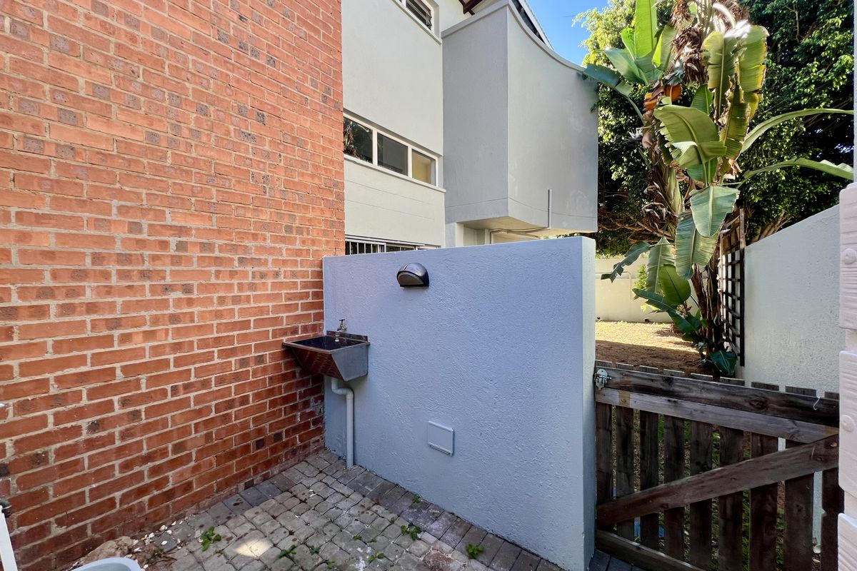 A functional rear exterior area featuring a brick wall, a small utility sink, paved flooring, and a wooden gate leading to the garden.