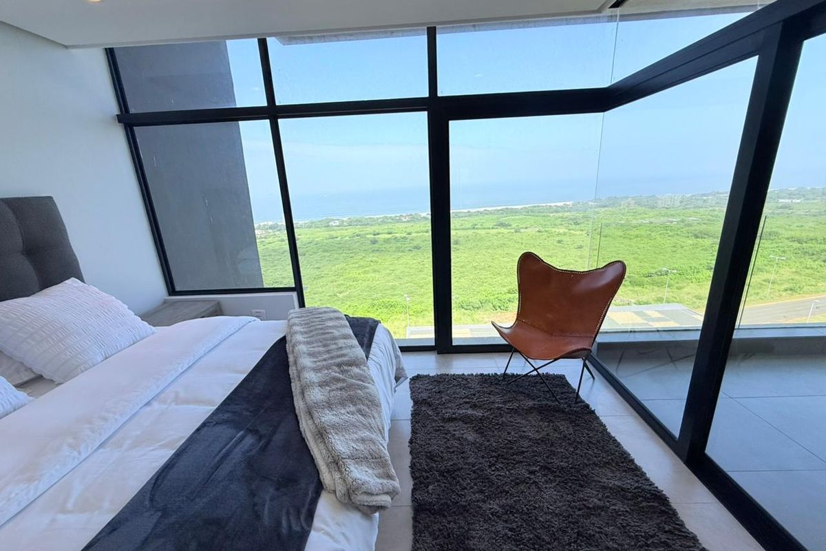 Modern bedroom featuring floor-to-ceiling glass windows with expansive coastal and landscape views, tiled flooring, and recessed ceiling lighting.