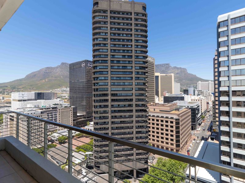 An elevated city view from a balcony, showcasing urban architecture and mountain scenery.