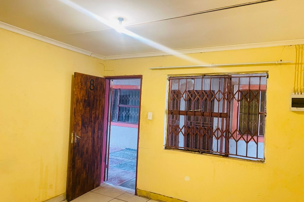 An empty room featuring tiled flooring, a wooden door, and a window fitted with a security gate, showing signs of wear and dated finishes.