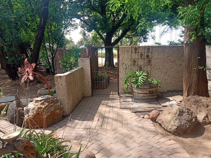 A landscaped garden area featuring a paved walkway, mature trees, and a secure boundary wall with a metal gate entrance.