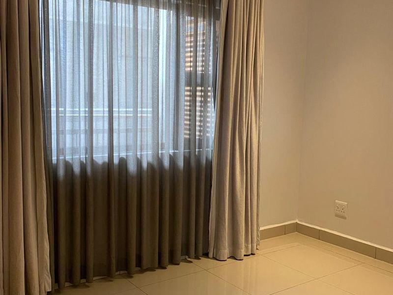 A bright, empty bedroom featuring neutral tiled flooring, recessed ceiling lighting, and floor-to-ceiling curtains over a large window.