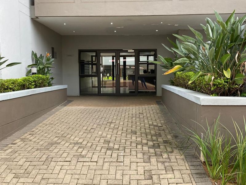 The entrance to the apartment building features a paved walkway flanked by raised garden beds and a covered entryway with glass doors.