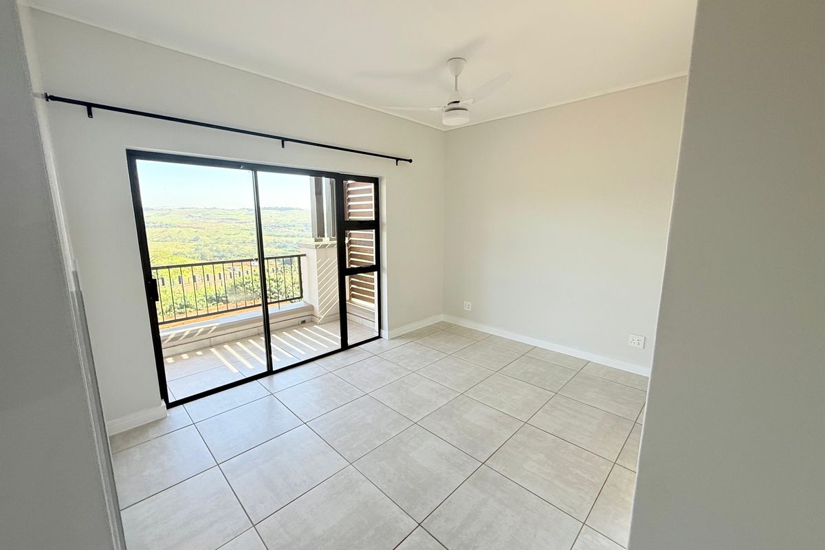 A bright, empty bedroom featuring neutral tiled flooring, a ceiling fan, and large sliding glass doors opening onto a private balcony with scenic views.