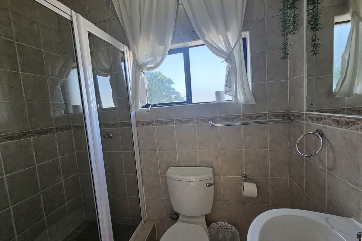 A functional bathroom featuring a shower enclosure, pedestal basin, and toilet with neutral wall tiling and natural light from a window.
