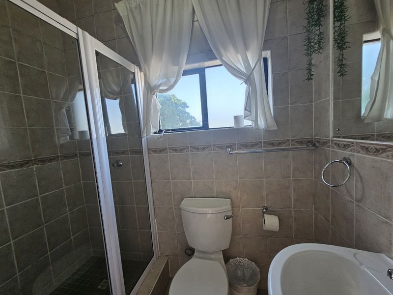 A functional bathroom featuring a shower enclosure, pedestal basin, and toilet with neutral wall tiling and natural light from a window.