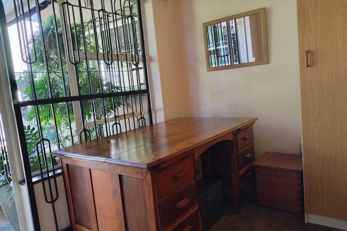 A bedroom featuring a built-in wooden wardrobe and a large window secured with metal burglar bars, overlooking a lush outdoor area.
