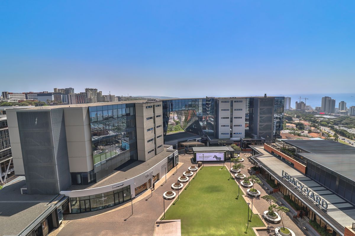 Aerial view of a modern commercial and residential complex featuring a central landscaped courtyard, glass-facade buildings, and surrounding urban infrastructure.