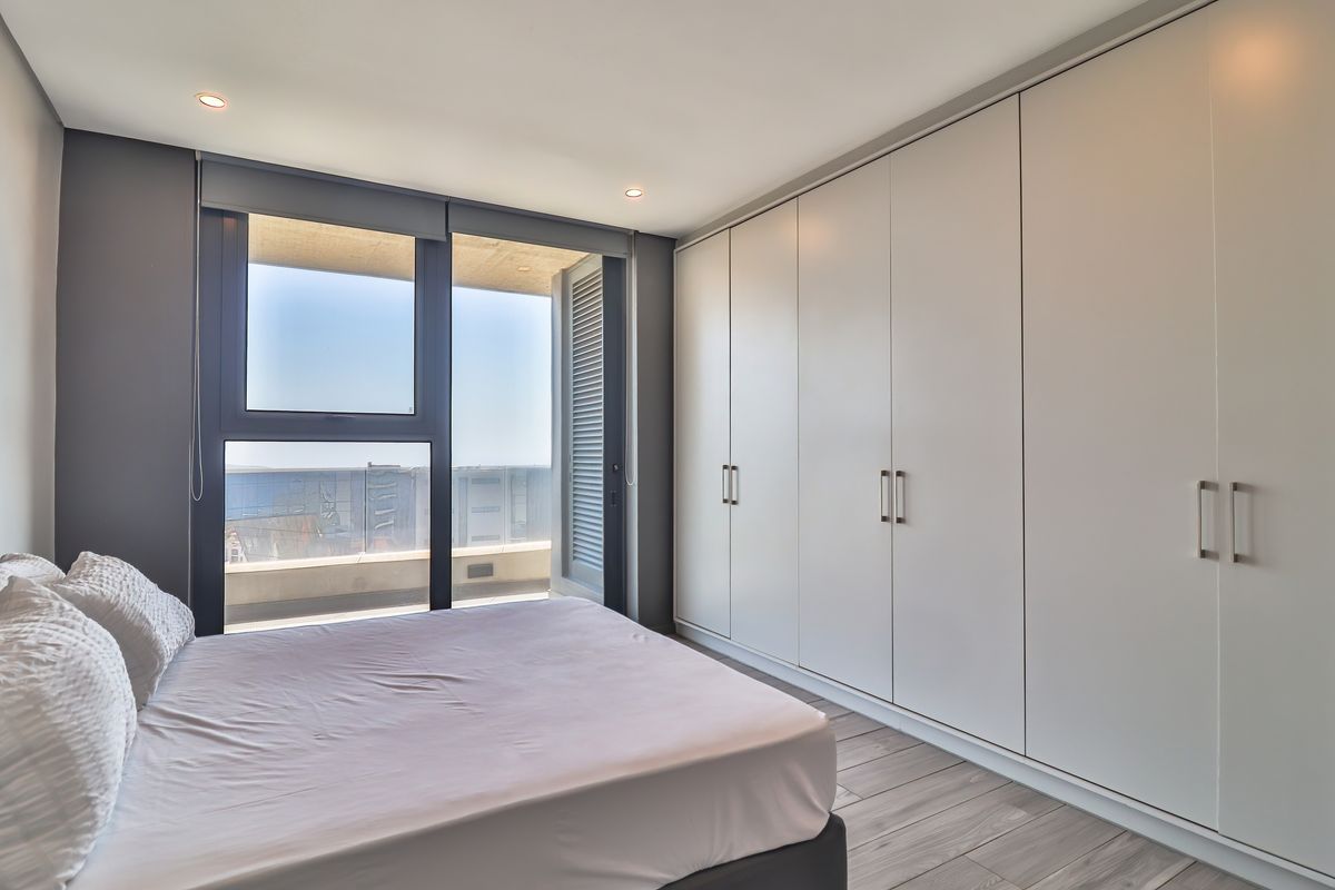 A bright bedroom featuring floor-to-ceiling built-in wardrobes, wood-look flooring, and large sliding glass doors opening to a private balcony with scenic views.