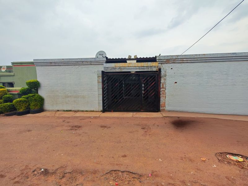 Front exterior view of the property featuring a painted boundary wall, metal security gate, and an unpaved driveway area.