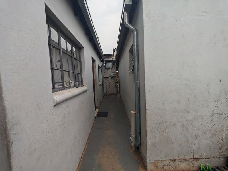 A narrow exterior side passage between two building structures featuring painted concrete flooring, metal-framed windows, and exposed downpipes.