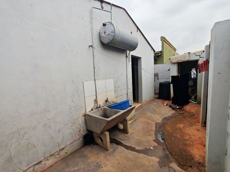 Exterior rear utility area featuring a concrete wash trough, wall-mounted geyser, and paved pathway leading to a secondary structure.