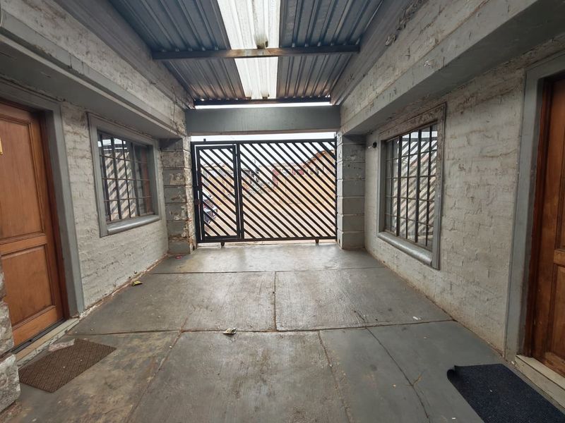 A covered outdoor entrance or carport area featuring a metal security gate, concrete flooring, and exposed brick walls.