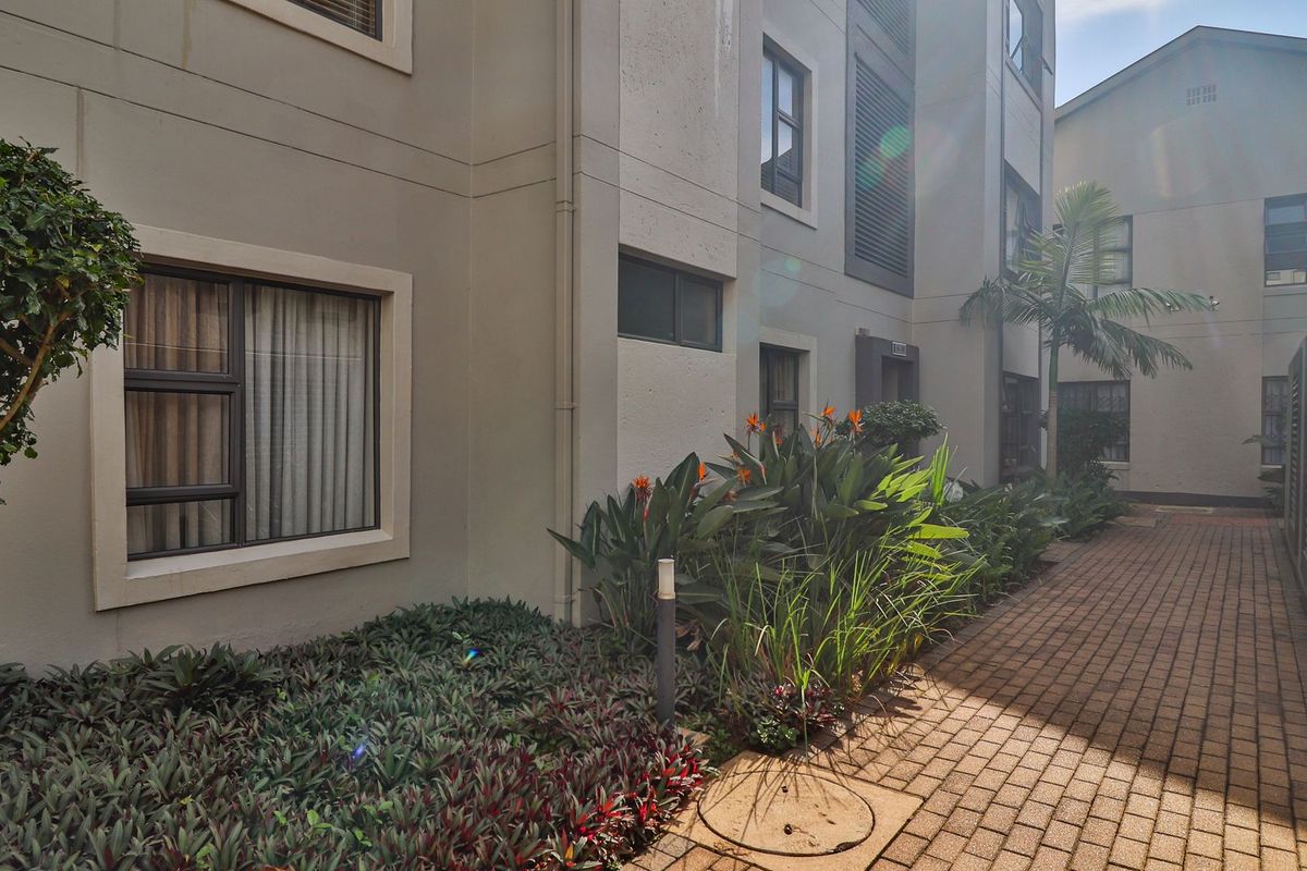A view of the apartment complex exterior featuring paved walkways, landscaped garden beds, and modern building facades with aluminium window frames.