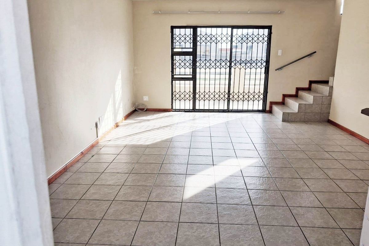 An empty, open-plan living area featuring tiled flooring, a staircase with a handrail, and a secure metal gate leading to the exterior.