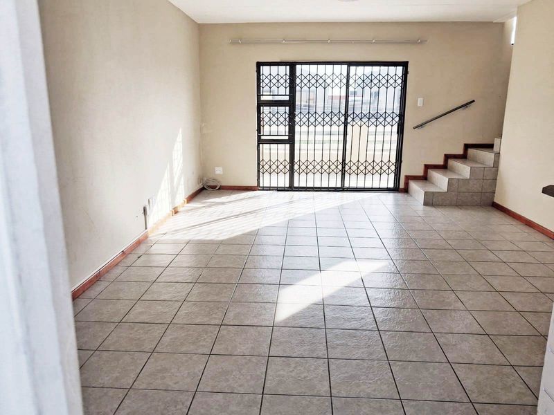 An empty, open-plan living area featuring tiled flooring, a staircase with a handrail, and a secure metal gate leading to the exterior.
