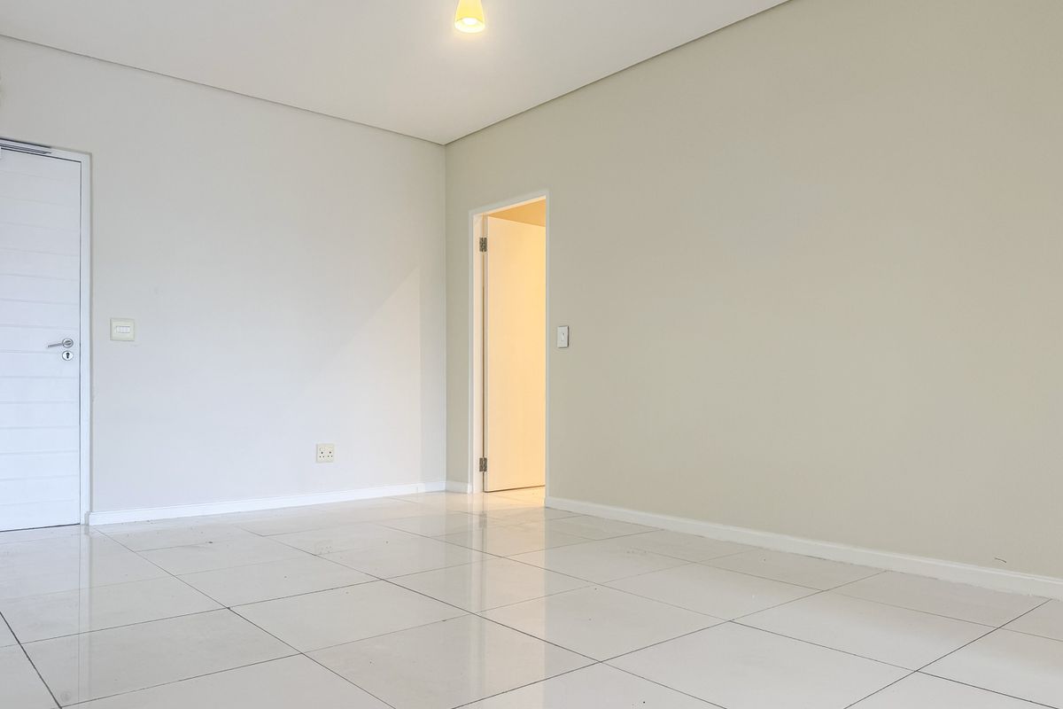 A bright, empty room featuring neutral walls, white tiled flooring, and a single pendant light fixture, leading into an adjacent space.