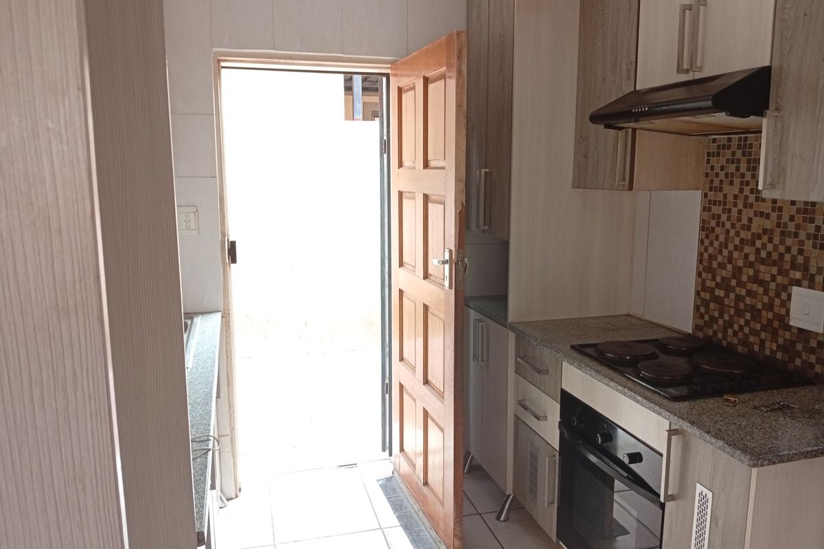 Compact kitchen featuring built-in cabinetry, granite countertops, mosaic tile backsplash, integrated oven, and hob with extractor fan, leading to an exterior doorway.