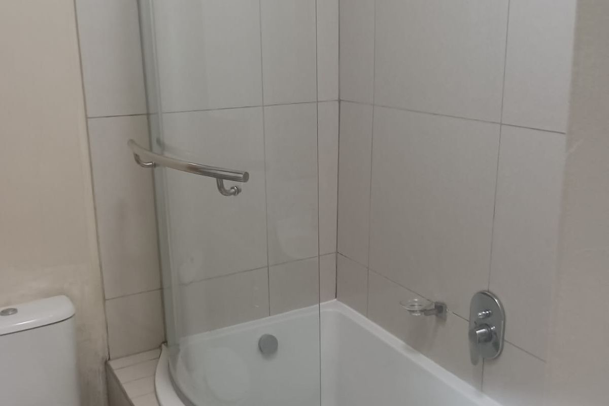 A functional bathroom featuring a curved shower-over-bath combination with a glass screen, tiled surround, and standard sanitary fixtures.