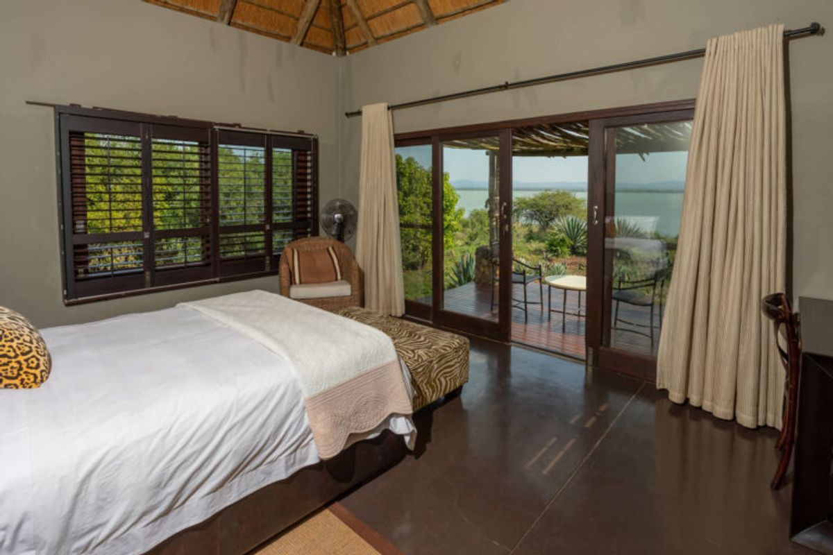 A bedroom featuring a high thatched ceiling, dark polished concrete flooring, wooden shutters, and sliding glass doors opening onto a private deck with water views.
