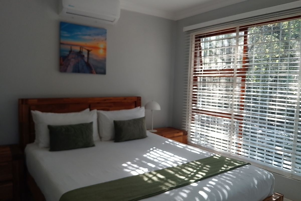 A well-maintained bedroom featuring a wall-mounted air conditioning unit and large windows fitted with horizontal blinds.