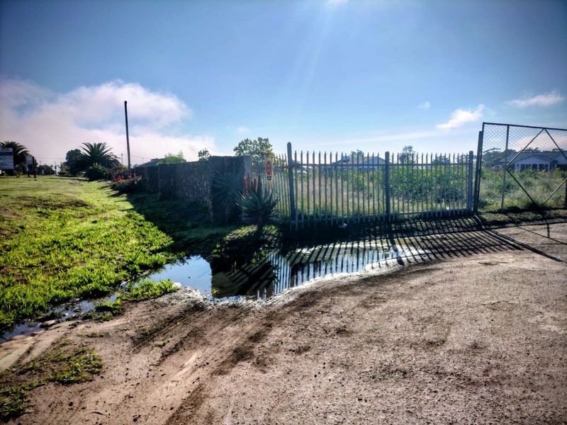 Vacant residential land featuring a metal security gate, stone boundary wall, and chain link fencing, with unpaved access and visible standing water.