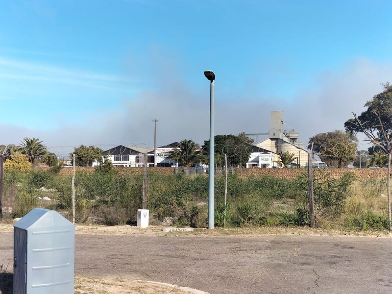 A street-level view of a vacant residential plot featuring perimeter fencing, a street light, and a utility box, with industrial buildings visible in the background.