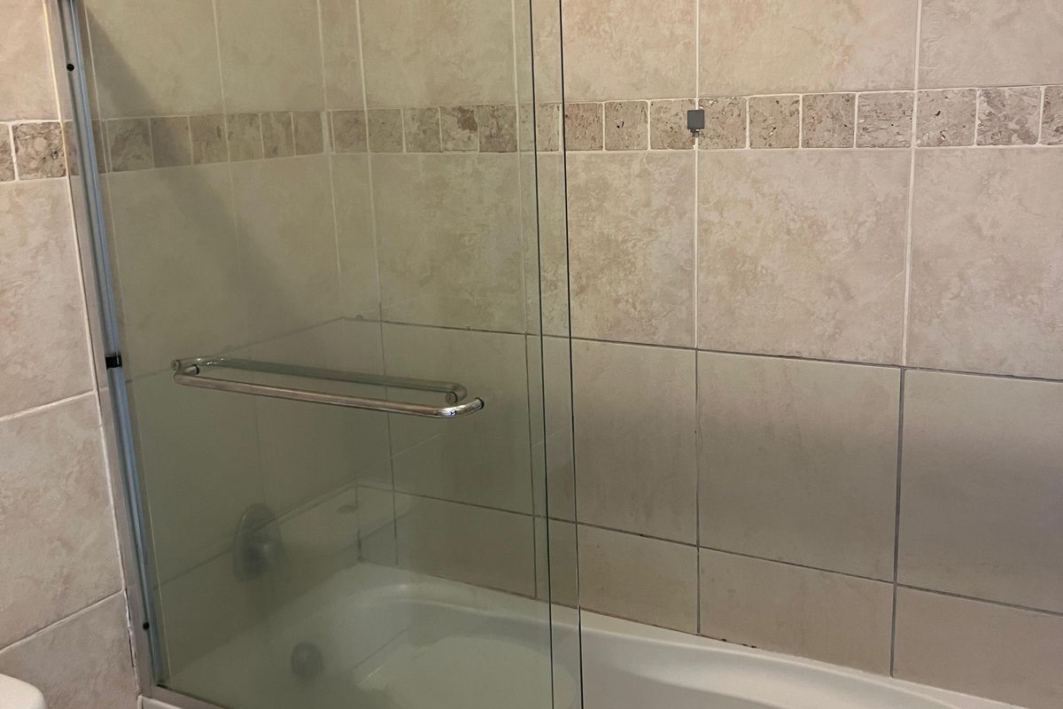 A functional bathroom featuring a built-in bathtub with a sliding glass shower screen and neutral wall tiling.