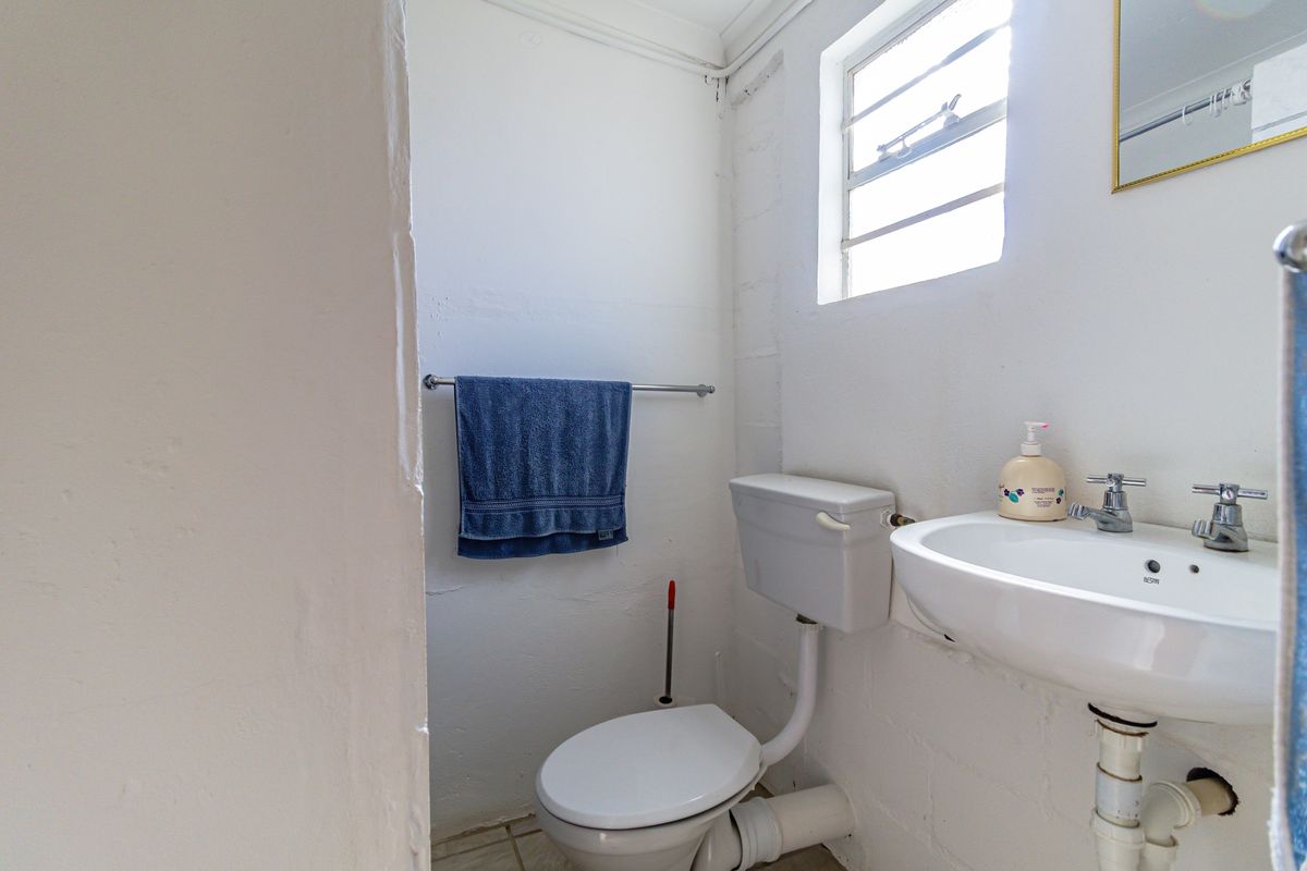 A compact bathroom featuring a standard toilet, ceramic basin, and louvered window with exposed plumbing fixtures.