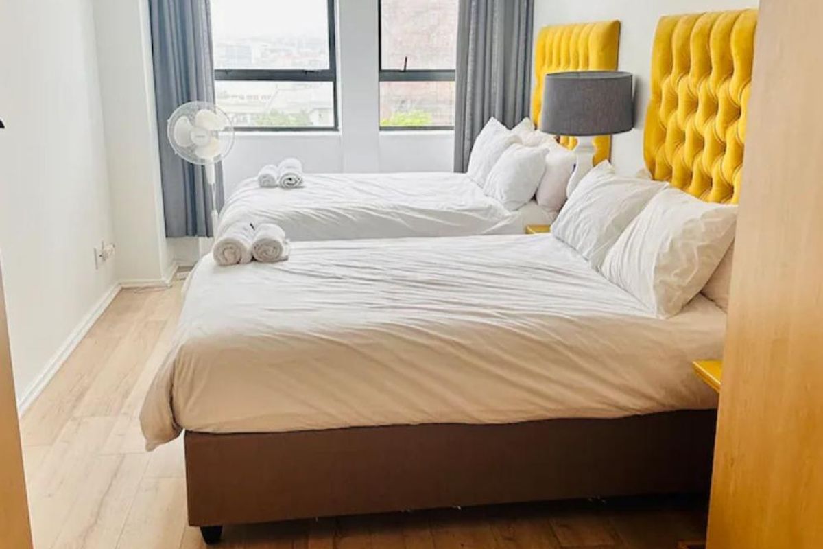 A bright bedroom featuring light wood-look flooring, large windows with city views, and neutral wall finishes.
