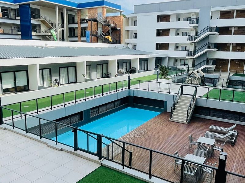 A well-maintained apartment complex courtyard featuring a central swimming pool, timber decking, artificial lawn areas, and modern glass balustrades.