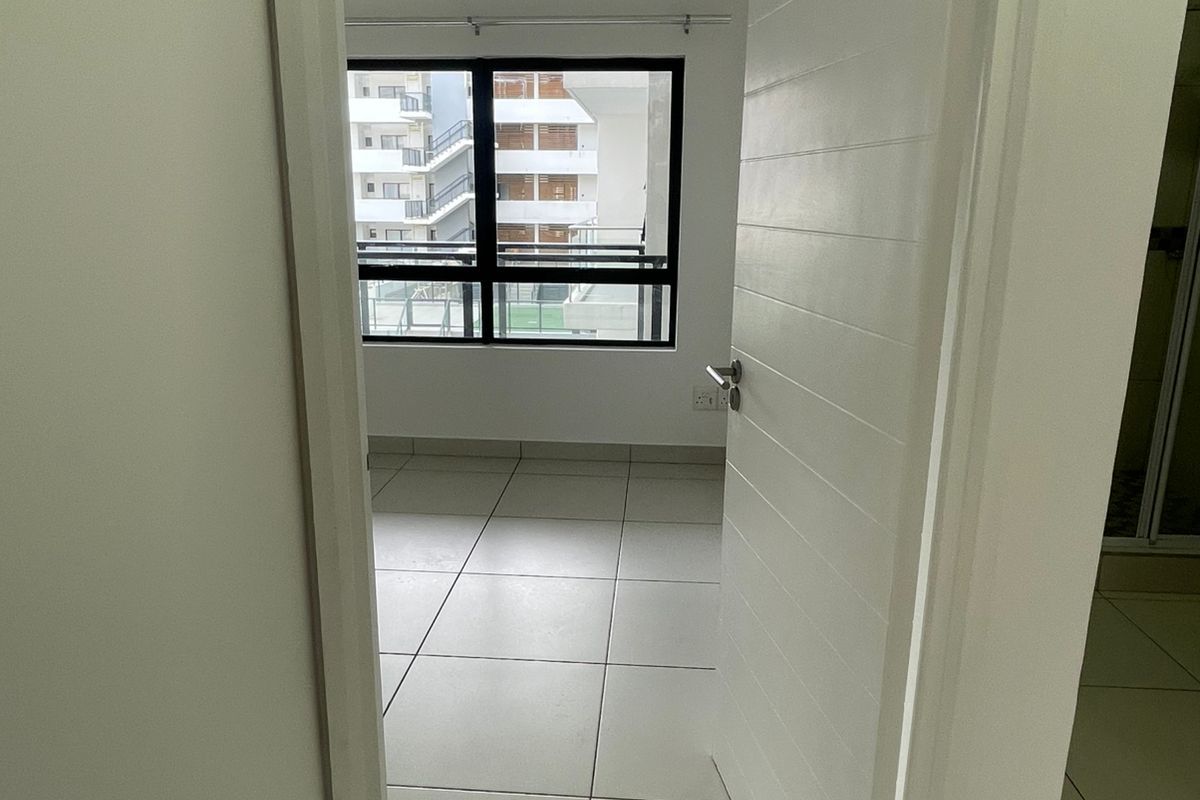 A view through a doorway into an empty bedroom featuring tiled flooring and a large window with a view of the surrounding apartment complex.