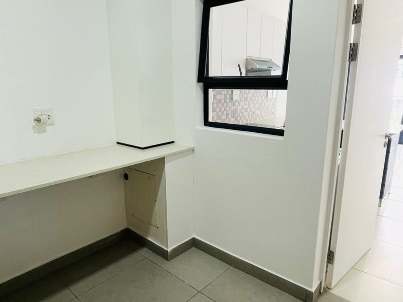A clean, functional laundry area featuring tiled flooring, a built-in countertop, and a window providing a view into the adjacent kitchen.