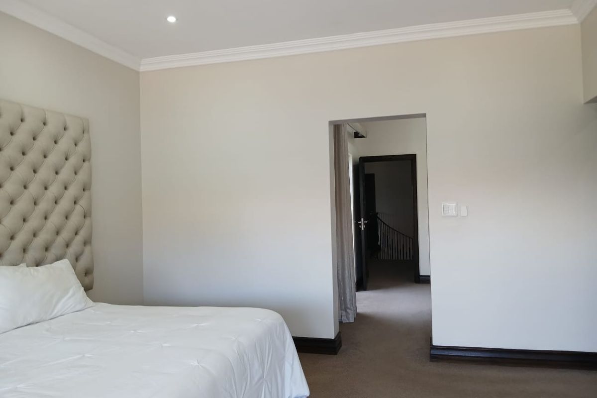 A spacious bedroom featuring neutral wall tones, recessed ceiling lighting, crown molding, and dark wood baseboards, leading into an adjacent hallway.