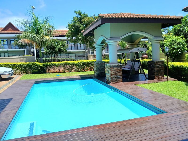 A well-maintained swimming pool area featuring a wooden deck surround and a covered gazebo with stone-clad pillars.