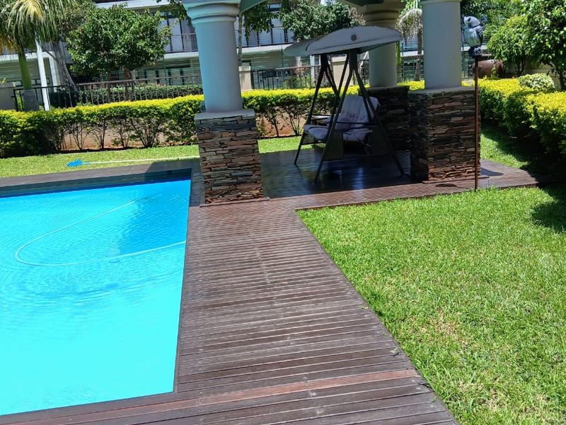 A well-maintained swimming pool area featuring a covered patio with stone-clad pillars and timber decking, set within a landscaped garden.