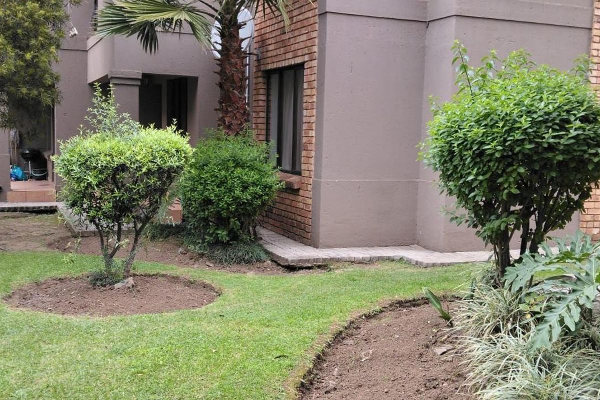 A view of the rear garden area featuring landscaped beds, established shrubs, and paved pathways adjacent to the apartment building exterior.