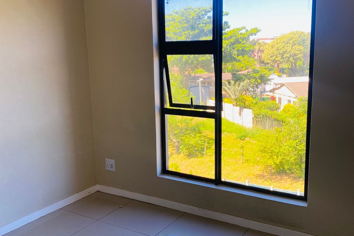 A bright, empty bedroom featuring neutral tiled flooring, a large window with a garden view, and a curtain rod installed.
