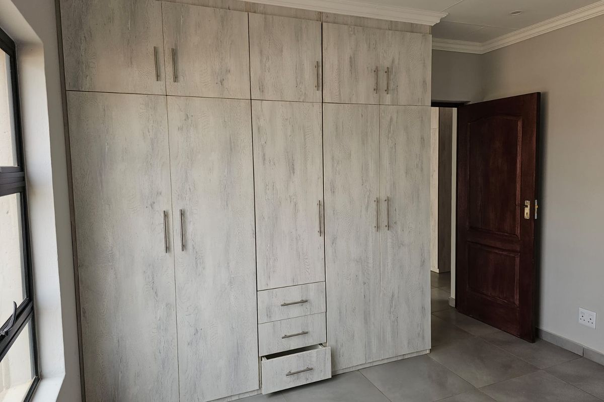 A bedroom featuring floor-to-ceiling built-in cupboards with a light wood-grain finish, tiled flooring, and a dark wood interior door.
