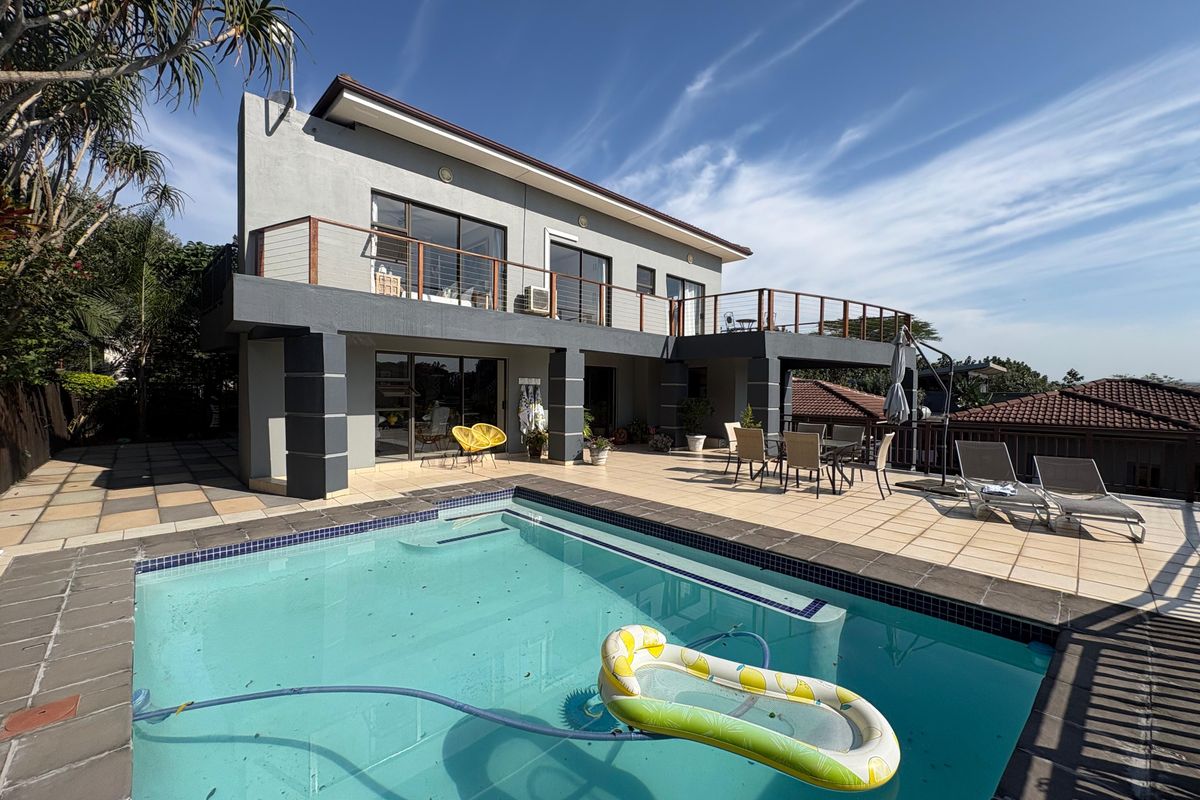 A spacious rear exterior view featuring a swimming pool, tiled patio area, and a two-story house with a balcony and sliding glass doors.