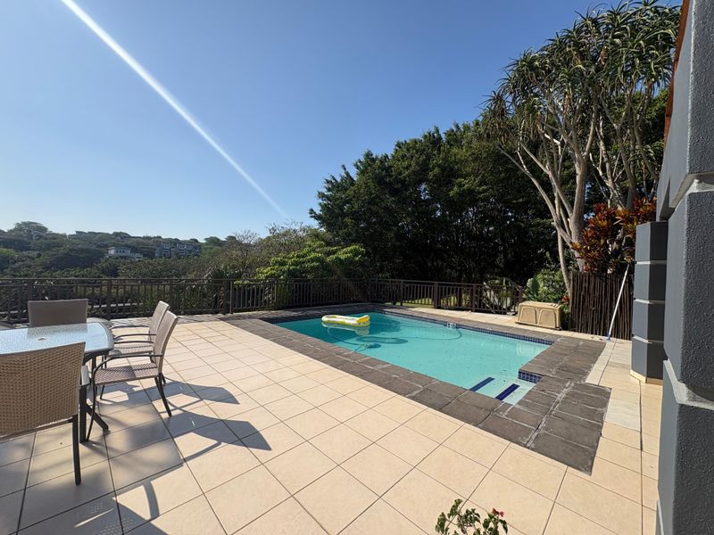 Outdoor pool area featuring a tiled patio, swimming pool with stone coping, and perimeter railing overlooking a lush green landscape.