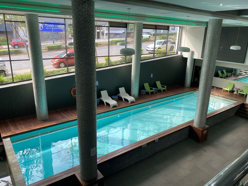 Indoor swimming pool area featuring mosaic-tiled structural columns, timber decking surrounds, and large floor-to-ceiling windows overlooking the street.
