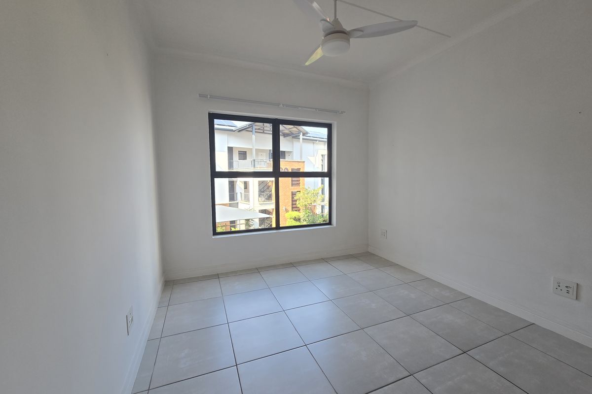 A bright, empty bedroom featuring neutral floor tiles, a ceiling fan, and a large window providing natural light and a view of the complex.