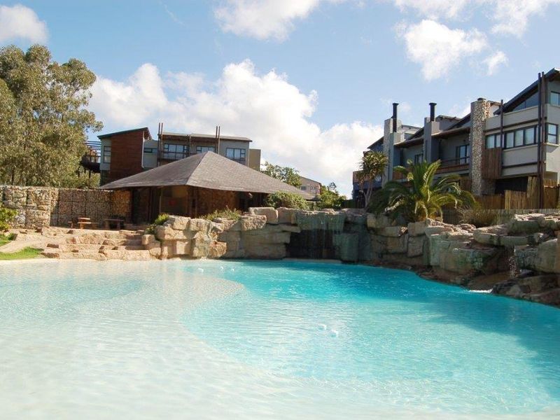 A well-maintained communal swimming pool area featuring a natural rock waterfall, beach-style entry, and a thatched gazebo set within a landscaped apartment complex.