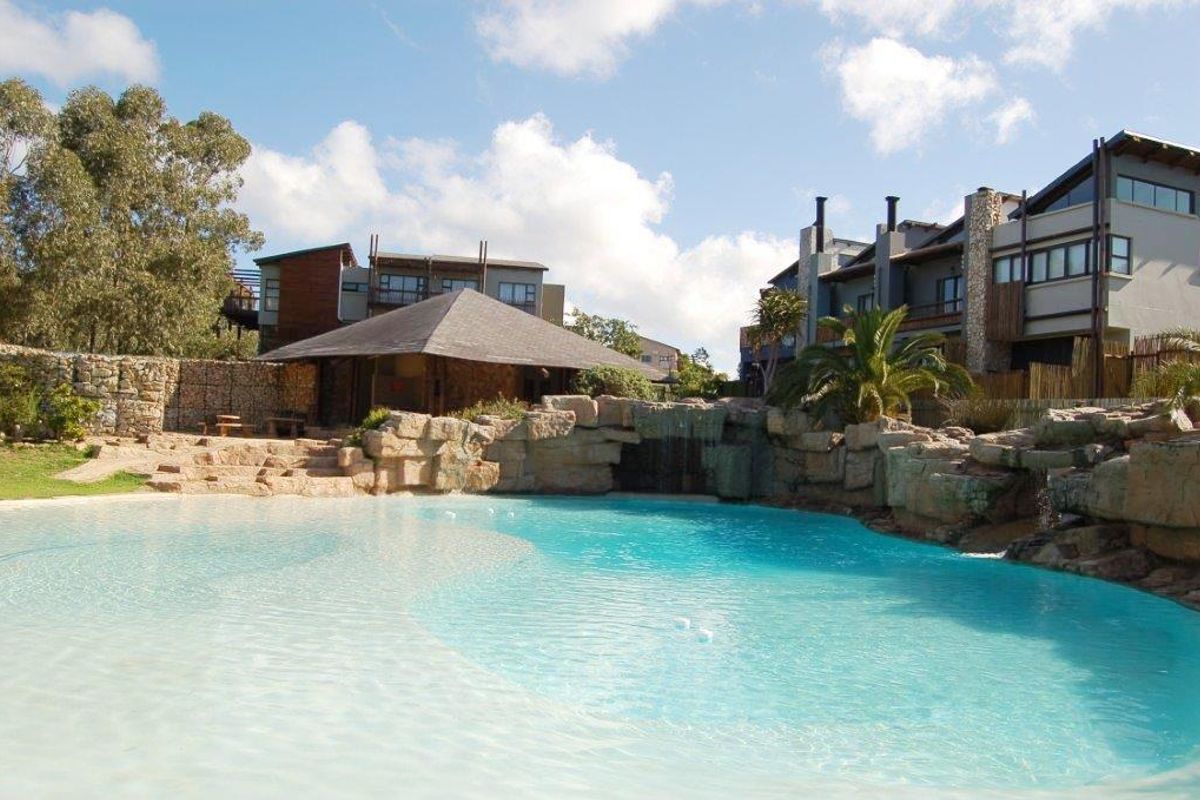 A well-maintained communal swimming pool area featuring a natural rock waterfall, beach-style entry, and a thatched gazebo set within a landscaped apartment complex.