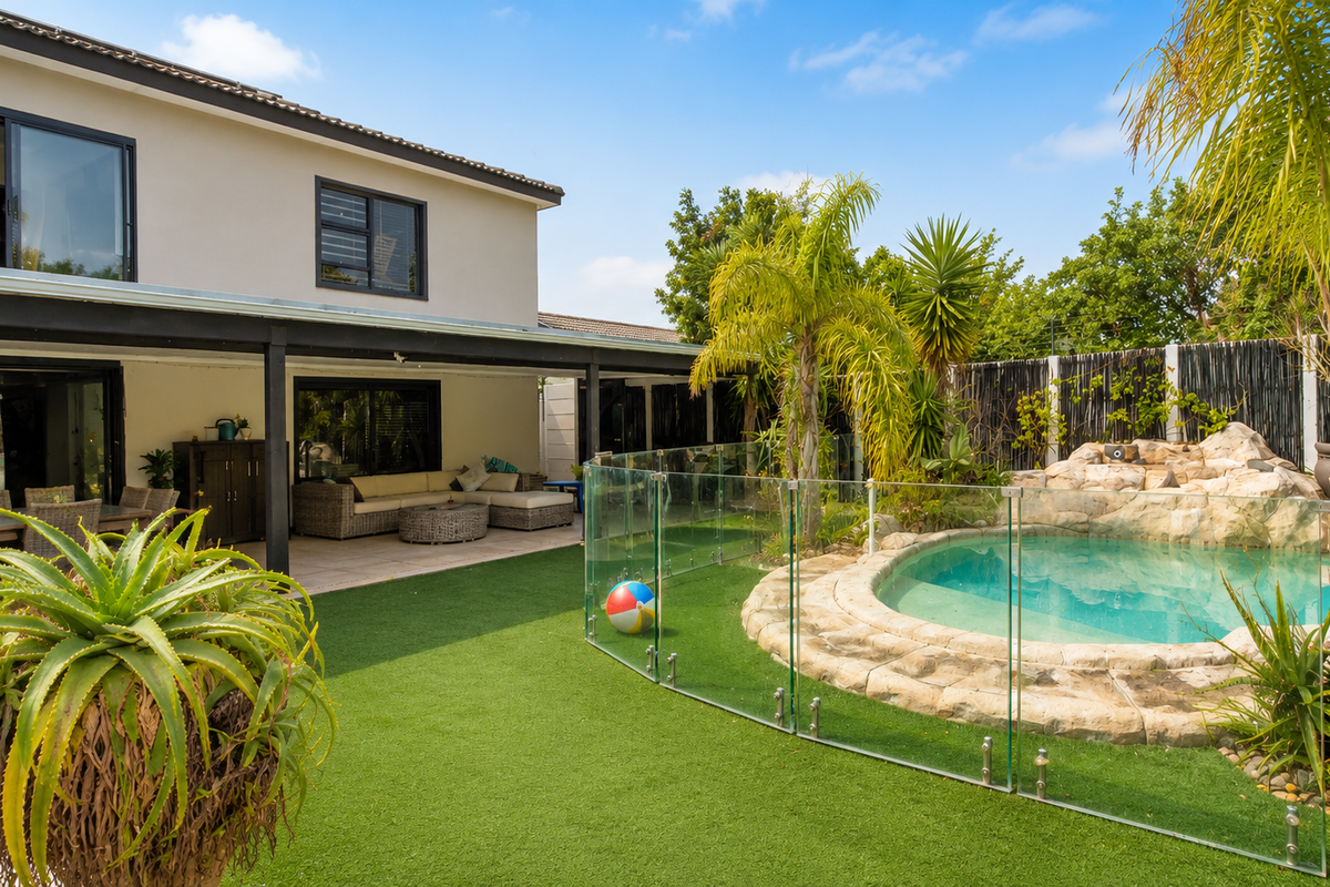 Spacious rear garden featuring a covered patio, artificial lawn, and a rock-feature swimming pool enclosed by a modern glass balustrade.
